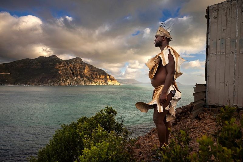 A man wearing tribal clothing looks out to sea. To his right is a small hut and in the background a vast mountain range. Taken by Brent Stirton on a Canon EOS R5.