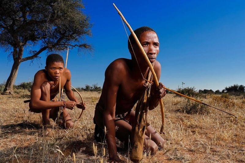 Two young men wearing tribal clothing and carrying handcrafted bows and arrows crouch down among yellowing grass. Taken by Brent Stirton on a Canon EOS R.