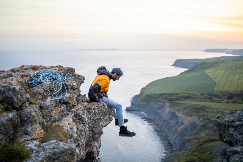 A man sits on the ledge of a cliff, his feet dangling over the edge, overlooking a sweeping coastline with craggy inlets snaking into the distance. 