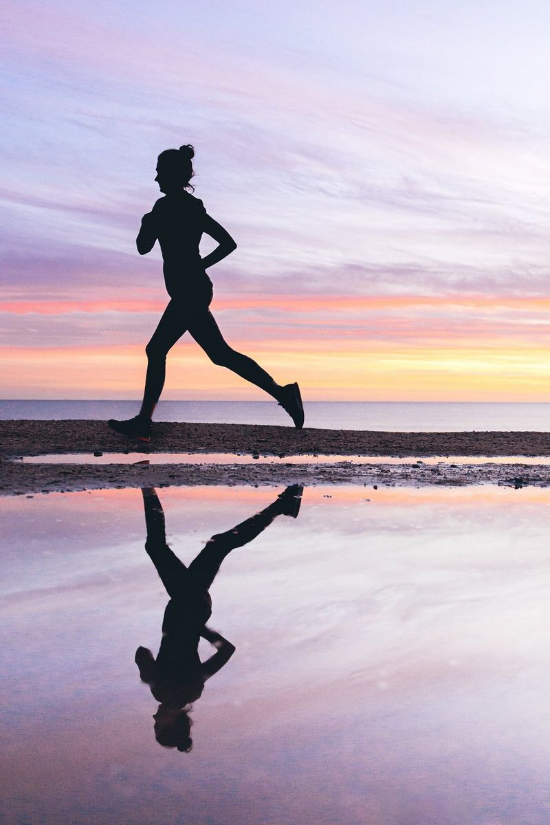 A runner in near silhouette is reflected in still water as she runs along the shore at sunset. 
