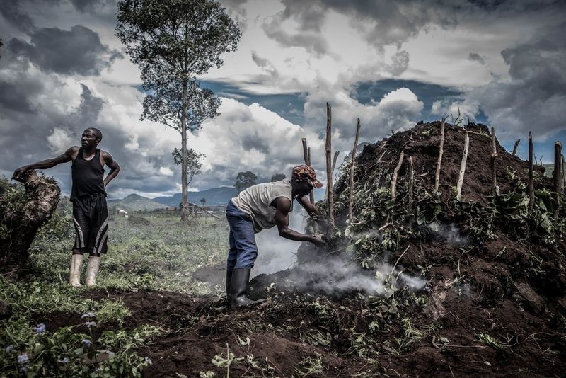 Two men in wellington boots beside a smoking kiln made with earth, one adding fuel. 