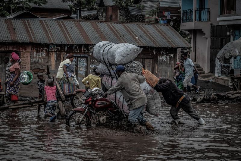 Two Congolese men struggling to push heavily laden motorcycles through flooded city streets. 