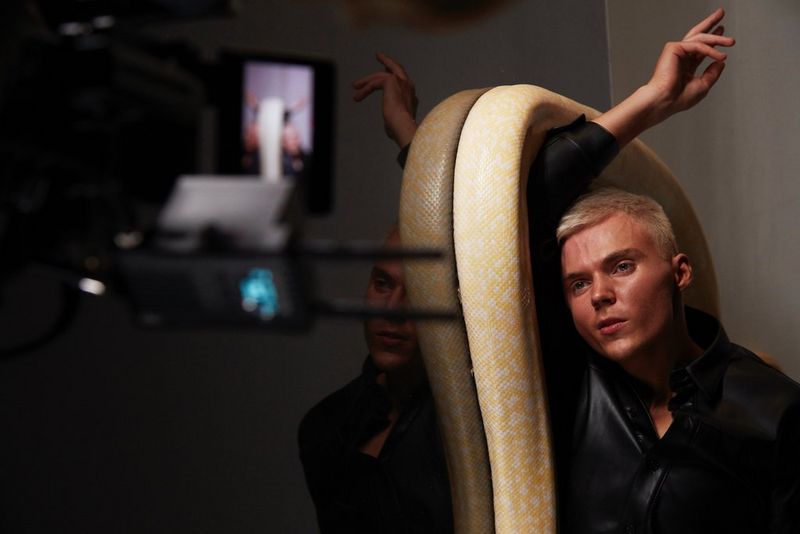 A Canon Cinema EOS camera films a young man with short blond hair lying on the floor. An enormous, yellow Burmese python is coiled around his raised arm.