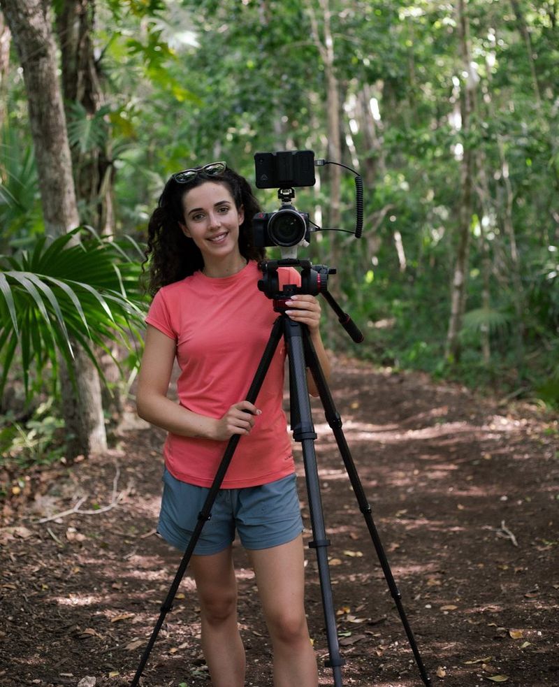 Filmmaker Roxy Furman stands in woodland, next to a Canon EOS R5 C camera on a tall tripod.