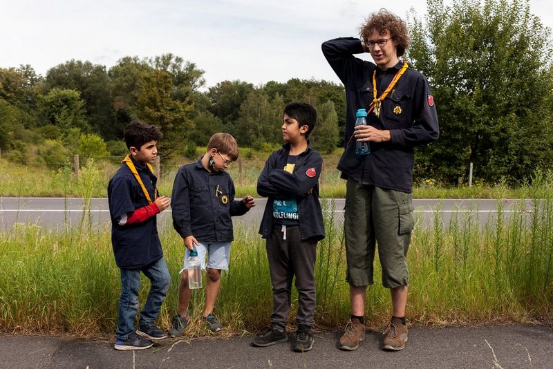 Three young scouts and one older scout leader stand waiting at the side of a country road. 