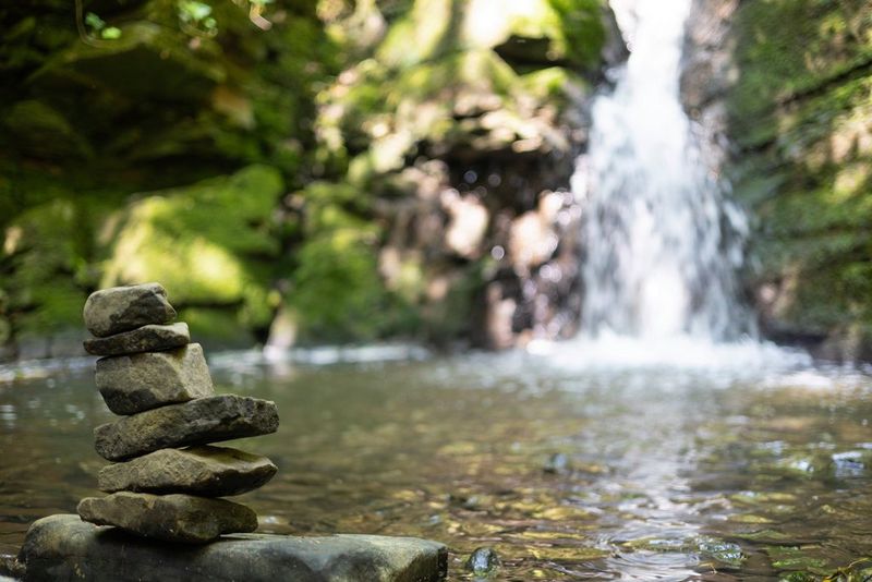 In a photo taken with a Canon RF 50mm F1.8 STM lens at f/1.8, a small stack of rocks stands in front of a pool of water, with a waterfall out of focus in the background.