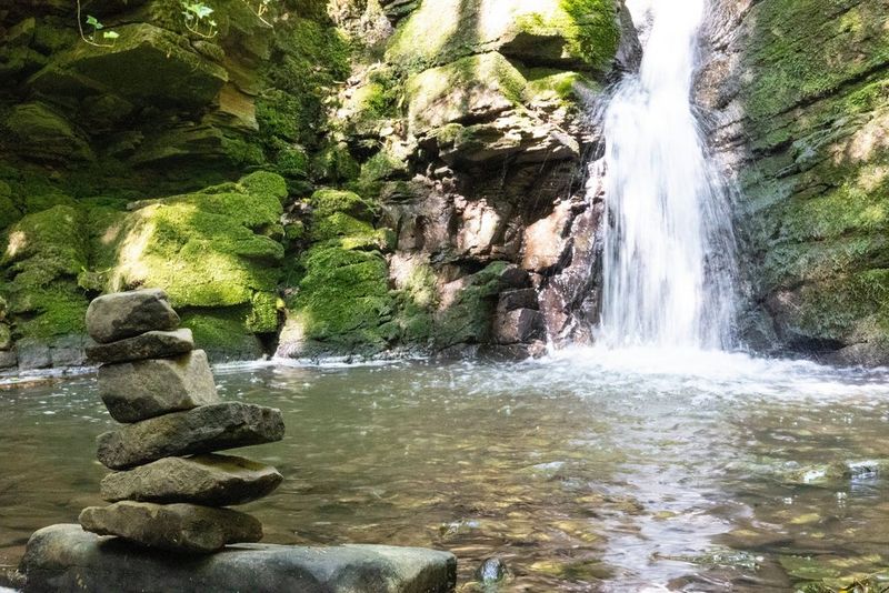 In a photo taken with a Canon RF 50mm F1.8 STM lens at f/22, a small stack of rocks stands in front of a pool of water, with a waterfall and rock face in the background all in focus.