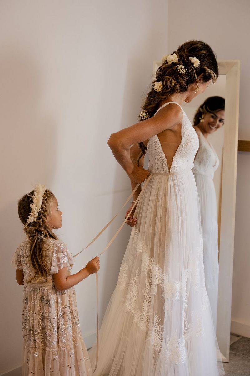 Head turned away from the camera and facing a full-length mirror, a bride adjusts the back of her dress with the help of her young bridesmaid.