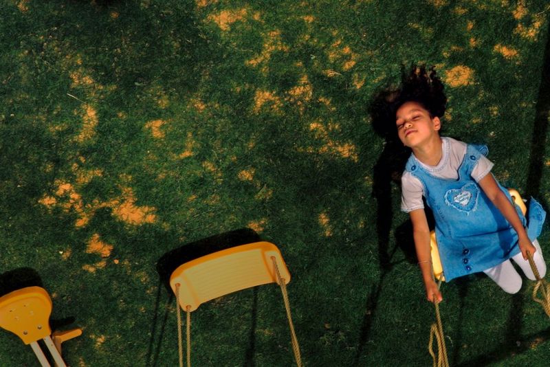 An image taken from above of a young girl in a denim dress happily playing on a swing. She is leaning backwards and her eyes are closed. 