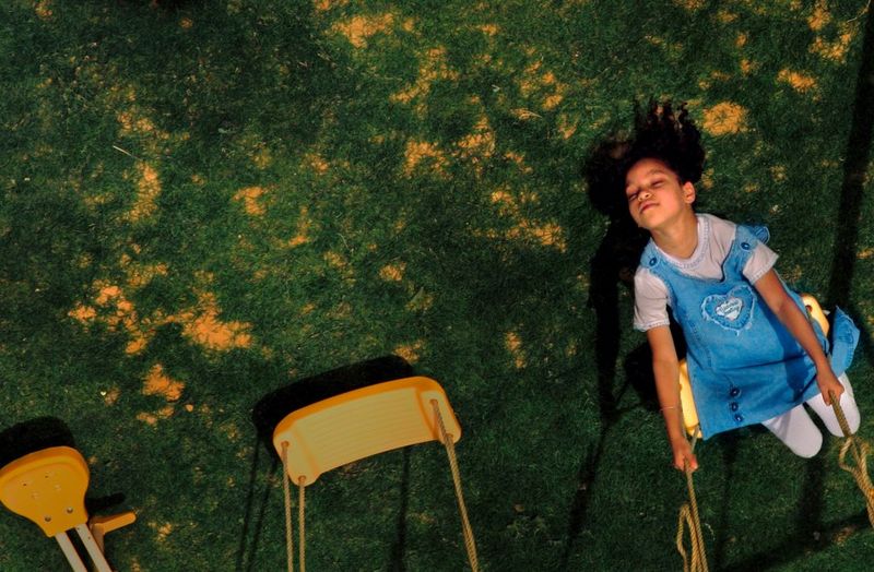 An image taken from above of a young girl in a denim dress happily playing on a swing. She is leaning backwards and her eyes are closed. 