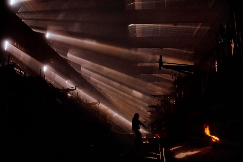 A person holds a hot steel rod, the end of which is glowing red. Only the silhouette of the person is visible as shafts of light shine through thick carcinogenic dust on the left side of the room. This photograph was captured by James Nachtwey in Czechoslovakia in 1990.