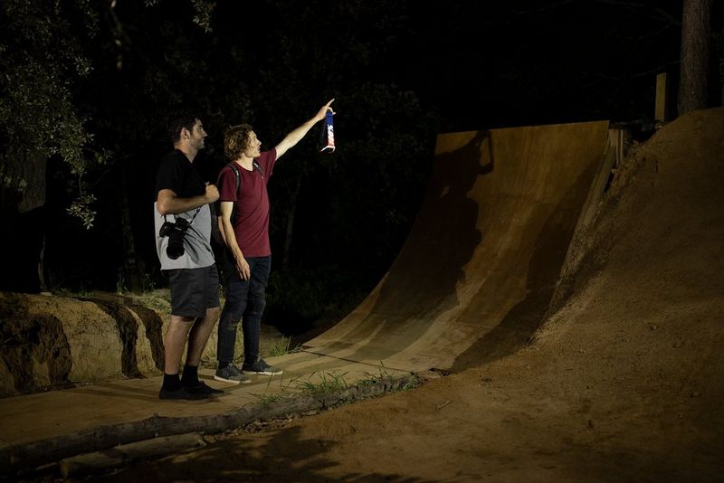 Two men standing at the bottom of a bike ramp at night. One of the men is pointing upwards, while the other has a Canon EOS R5 camera around his neck. 