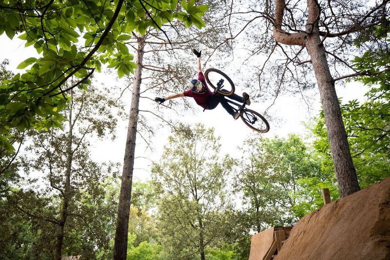 A bike rider performs a daring jump in a forest. His arms are aloft and he is at least several metres off the ground. 