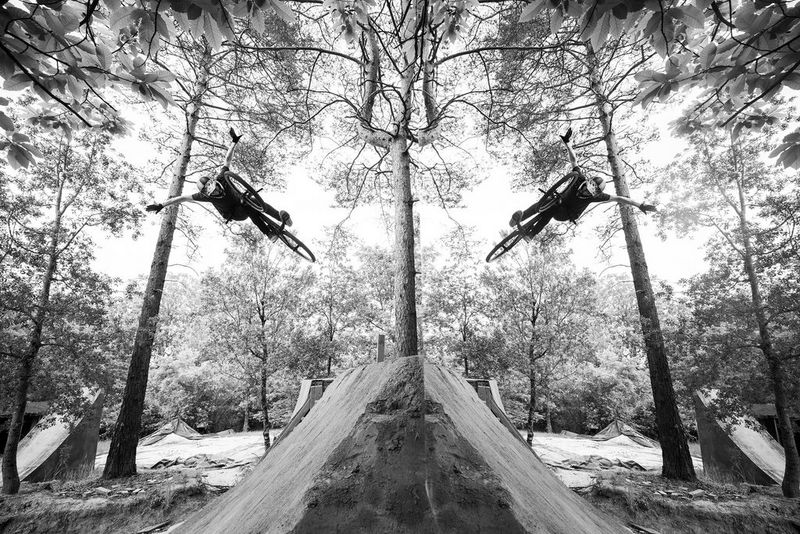 A black and white mirror image of a rider making a daring leap with his arms held aloft in a forest. 