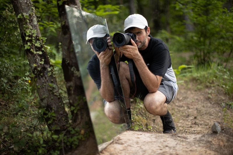 A photographer crouches in a forest taking a picture with a Canon EOS R5 camera. Next to him is a large mirror.
