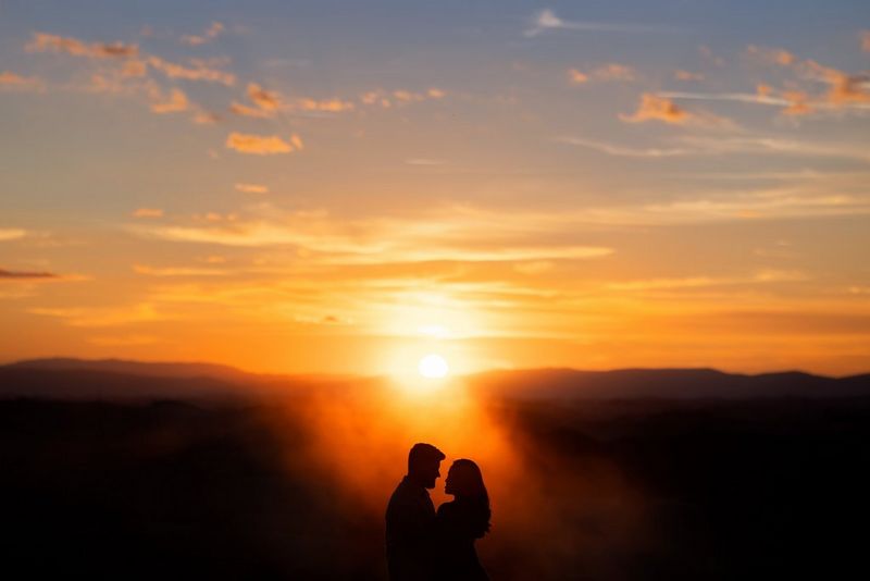 A couple embracing silhouetted against a dark mountain range and lit by the sun setting below an orange, cloud-streaked sky.