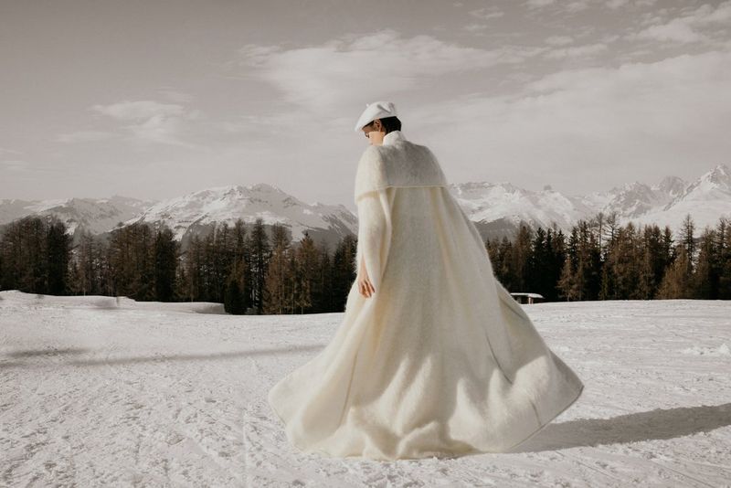A woman in a white hat and a long white coat walks away from the camera across a snowy landscape.