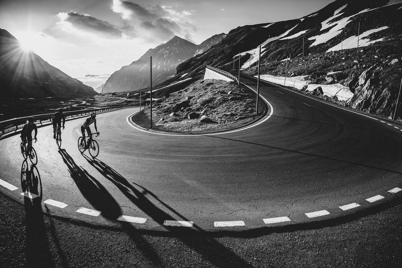 A black and white image of three cyclists approaching a hairpin bend on a mountain road. The low sun behind them casts long shadows onto the road surface.
