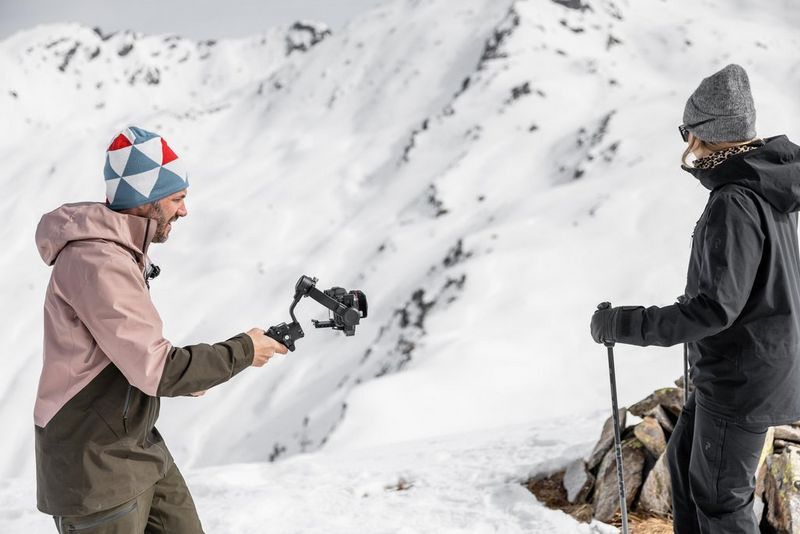 A man holds out a gimbal with a camera on the end, filming a woman holding ski poles, against a mountainous snowy background.