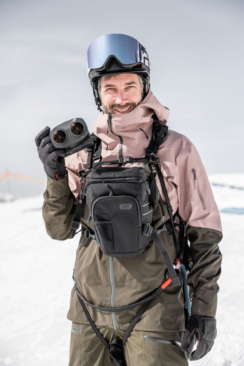 A man in outdoor clothing and helmet with a camera bag strapped to his front holds up a camera with dual fisheye lens.