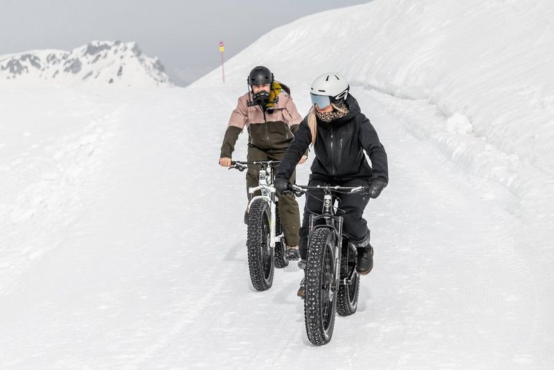 Two people on mountain bikes ride through the snow. The one at the back is wearing a helmet with a camera rigged on the front.
