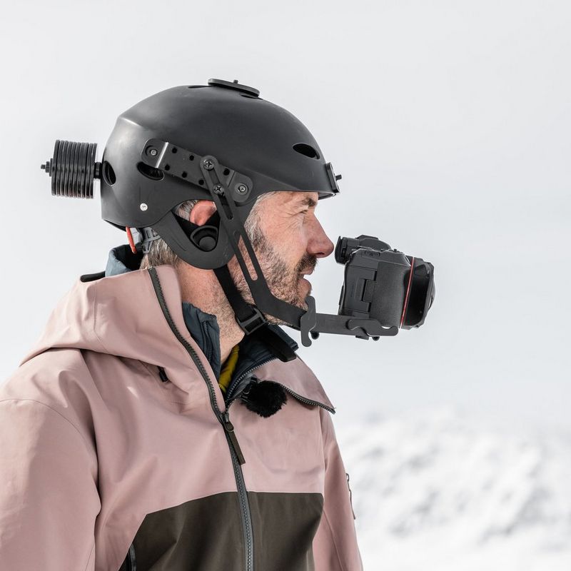 A man stands against a snowy background and white sky, wearing a helmet with a camera rigged to the front and counterweights on the back.