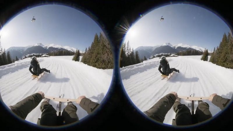 Two side-by-side fisheye images of a person sledding downhill, with snow-covered mountains in the distance. The filmmaker's handlebars and feet are visible in the foreground.  