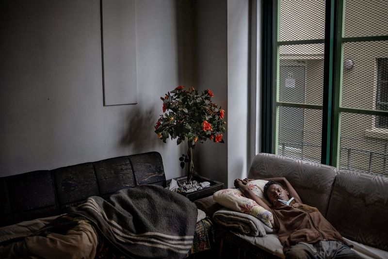 A woman lies across a sofa with her hands draped above her head, a face mask pulled down to her chin. In the corner of the grey room is a small planted tree with bright orange flowers.