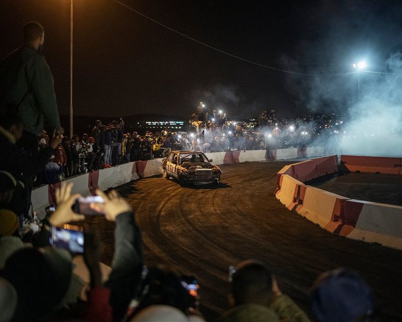 A car skids around a track at night, with spectators lining the outside of the track taking photographs.