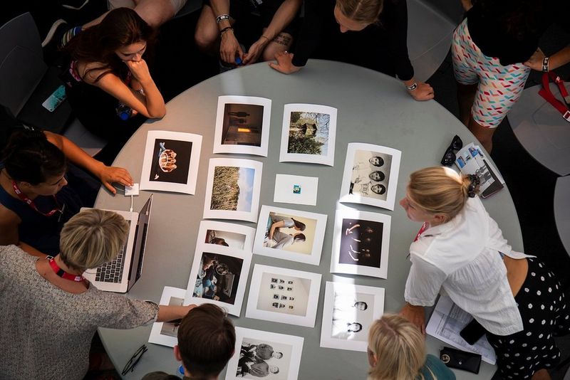 A group of people sit around a round table, which is laid out with photo prints.