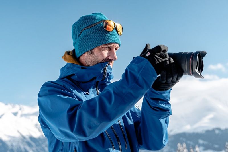A man, wearing a blue coat and woolly hat, looks at the screen on the back of a Canon EOS R5 camera. Snow-capped mountains can be seen blurred in the background behind him.