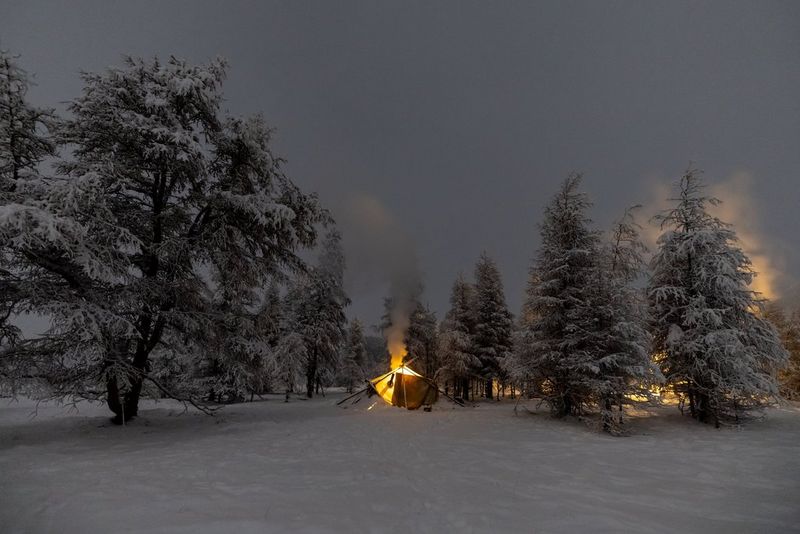 A tent pitched up on a snowy landscape, surrounded by trees covered in snow, while a campfire burns behind the tent, captured on a Canon EOS R3. 