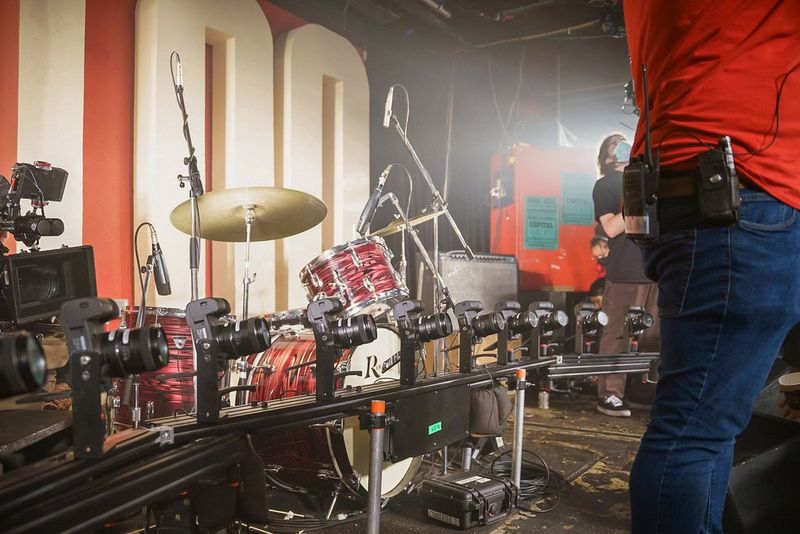 Crew members adjust cameras and props on a stage containing a bar cam, an electric guitar and an elaborate drum kit. In the background, the number 100 can be seen.