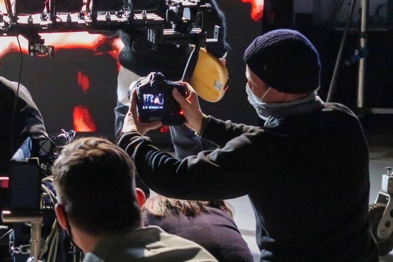 A masked crewmember films with a Canon EOS-1D X Mark III camera on a dark television set. Several other crewmembers watch on.
