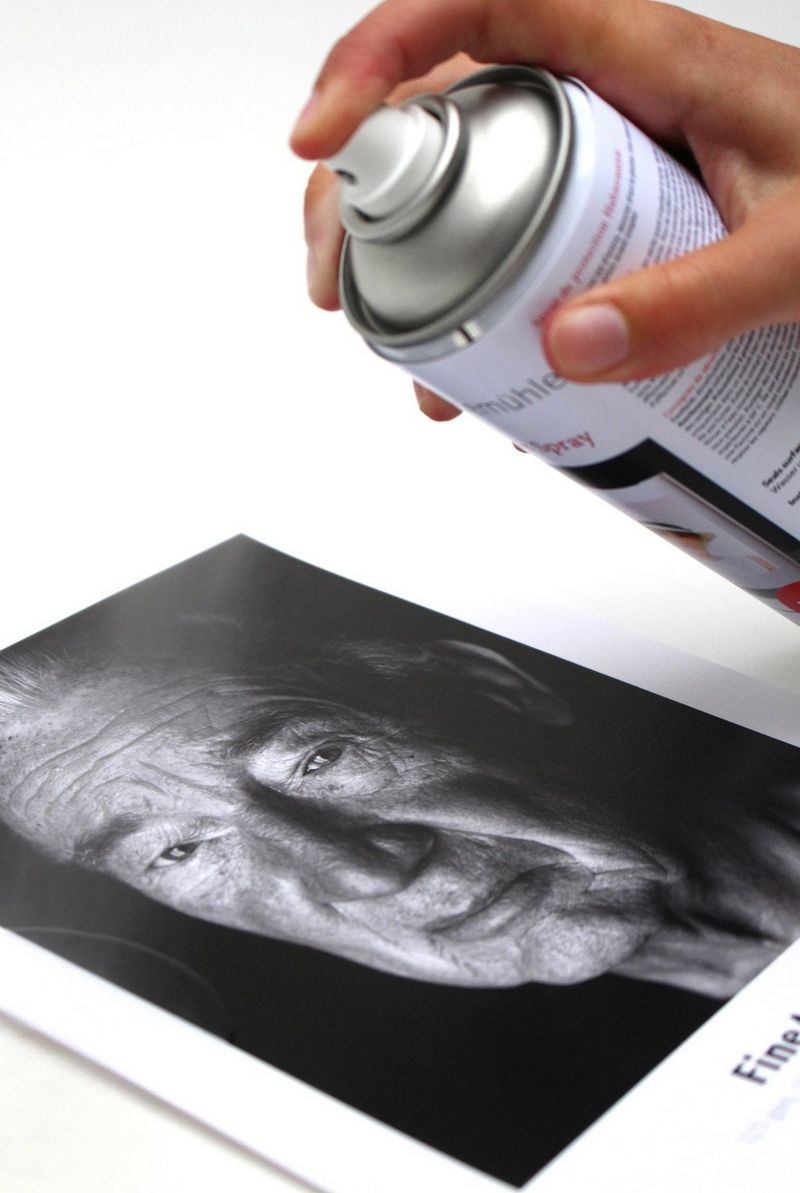 A hand holding a can of Hahnemühle Protective Spray over a black and white print of the face of an older man. 