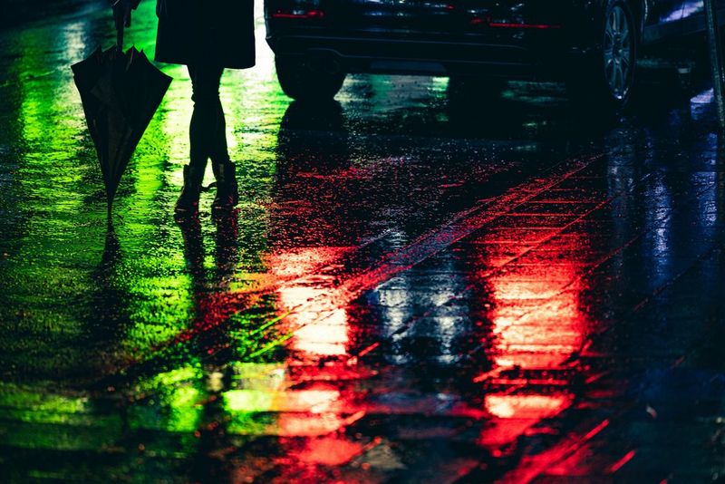 Green and red lights reflected in the puddles on a wet road with a woman holding an umbrella in silhouette. Taken on a Canon EOS R6 with a Canon RF 70-200mm F2.8L IS USM lens.