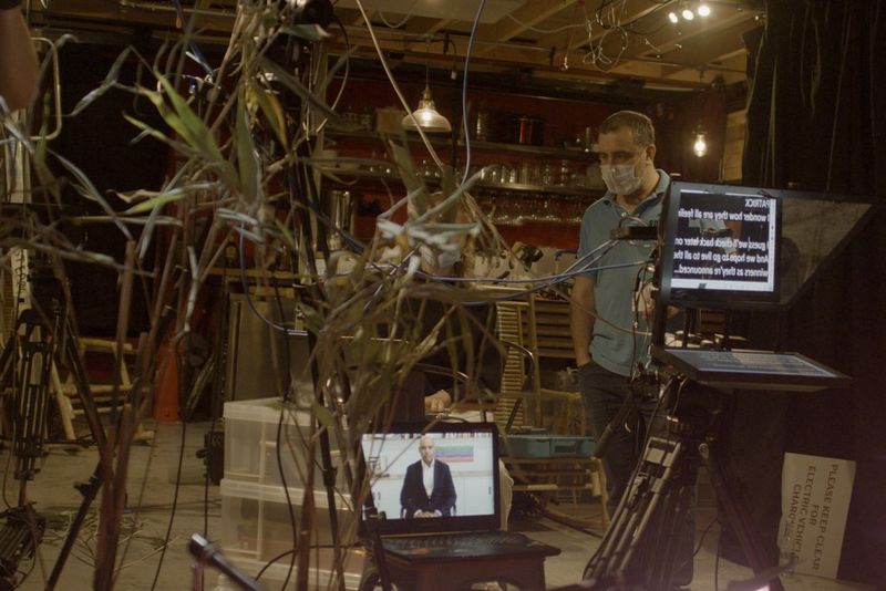 A man on a makeshift production set standing behind an autocue, with dresser shelves in the background and leafy plants in the foreground.