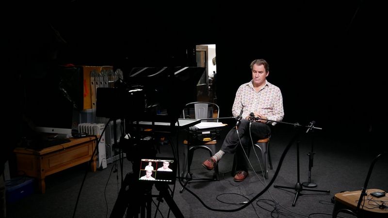 A man sits alone in a darkened studio with multiple views of him visible on the monitor attached to the camera in front of him.