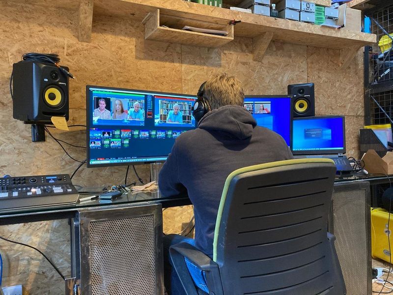 A man wearing headphones working on a three-monitor setup, editing footage of high-profile interviewees.