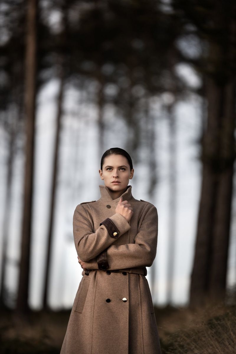 A woman in a brown coat with her arms wrapped around herself stands in a forest with trees blurred in the background. Taken by Sascha Hüttenhain with a Canon RF 135mm F1.8L IS USM lens.