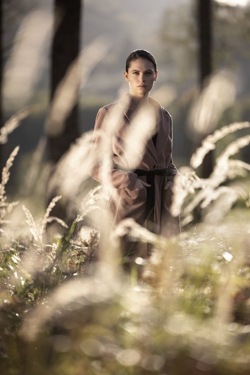 A woman in a brown coat with her hands in her pockets stands in a forest with wild grasses blurred in the foreground, while she looks directly at the camera. Taken by Sascha Hüttenhain with a Canon RF 135mm F1.8L IS USM lens.