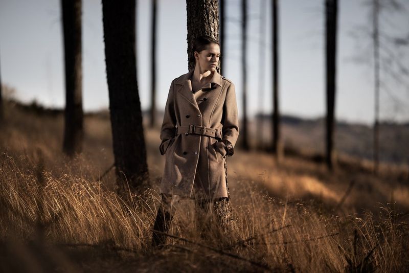 A woman in a brown coat with her hands in her pockets stands against a tree in a forest. Taken by Sascha Hüttenhain with a Canon RF 135mm F1.8L IS USM lens.
