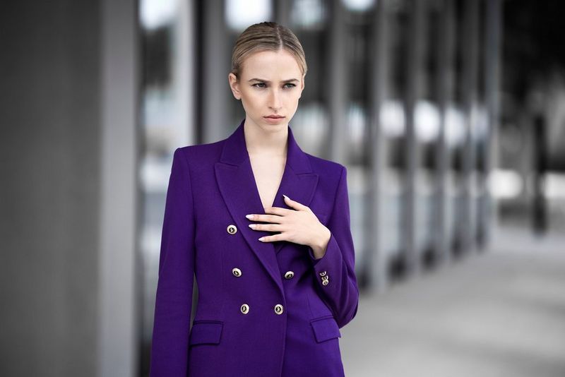 A woman in a purple blazer holds her hand to her chest and stands against a grey architectural background. Taken by Sascha Hüttenhain with a Canon RF 135mm F1.8L IS USM lens.