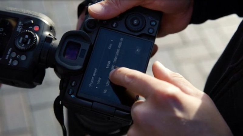 A photographer's finger adjusts shooting settings on the touchscreen of a Canon EOS R5 with a Speedlite EL-1 in its hot-shoe.