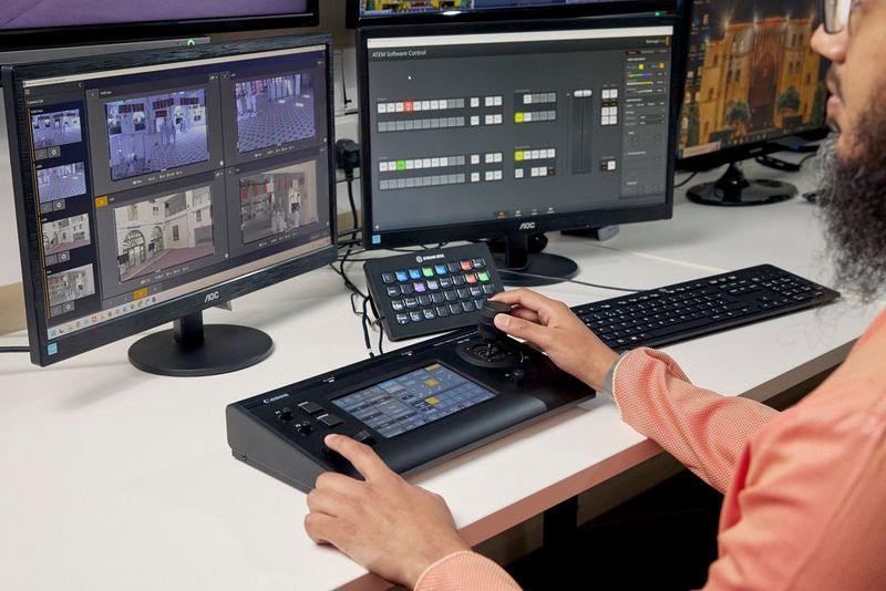 A user operating a Canon RC-IP100 hardware controller on a desk in front of a monitor showing the feed from multiple cameras.