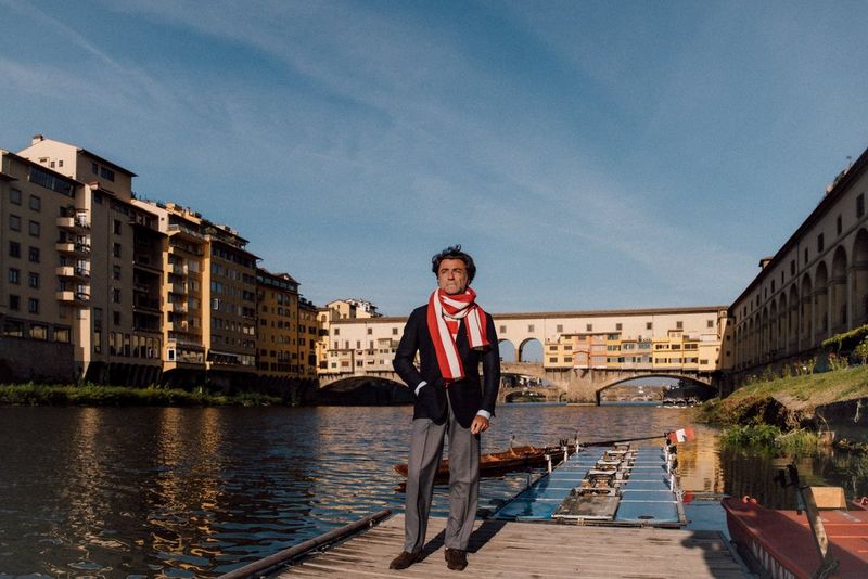 In a photo taken at a focal length of 24mm, a man in a blazer and scarf stands on a raft at the side of the river Arno in Florence, with the Ponte Vecchio in the background.