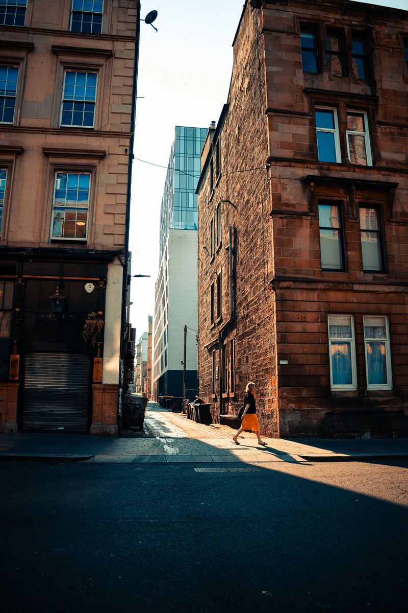 A figure caught in the sunlight between two tall buildings casts a long shadow on the pavement. Taken by Andres McNeill with a Canon RF 28mm F2.8 STM lens.