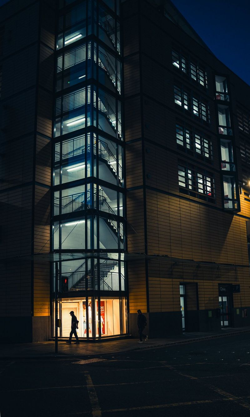 A person is illuminated by the ground floor lights of a multi-story carpark. Floor-to-ceiling windows show a staircase ascending to the top of the building. Taken by Andres McNeill with a Canon RF 28mm F2.8 STM lens.