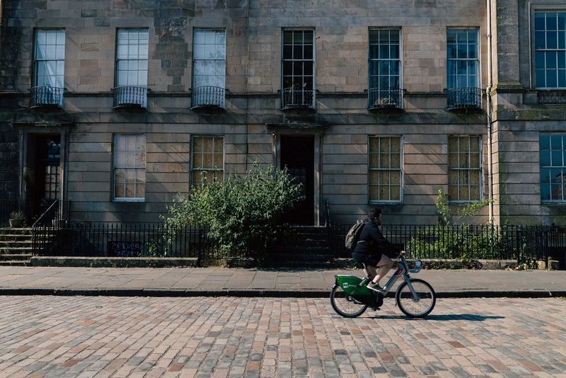 In a street photograph by Andres McNeill, a cyclist rides a bike down a cobbled street in front of a magnificent stone building.