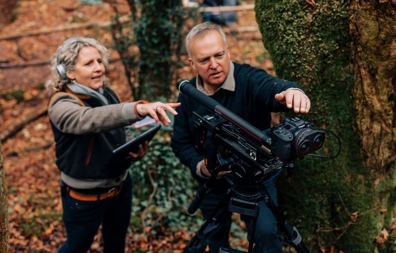 Two people in a wooded area gesturing at a camera on a stand with a Canon lens attached. 